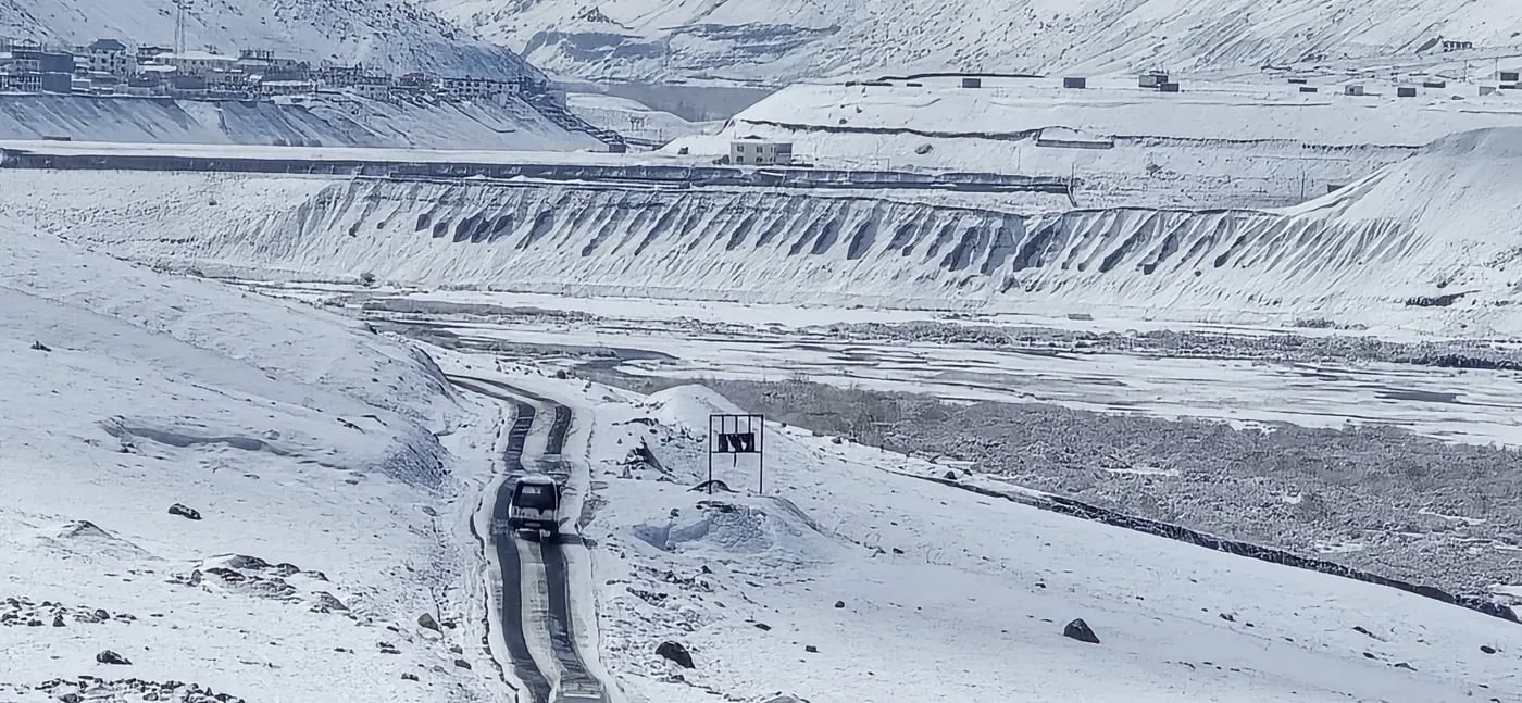 A car drives a narrow cleared road through snow in Spiti, with the river and white mountains beyond