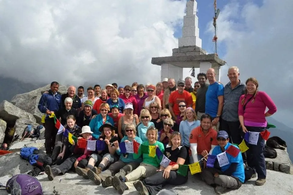A large group of trekkers, many in their sixties, sits with prayer flags beside a white stone shrine high in the Dhauladhar