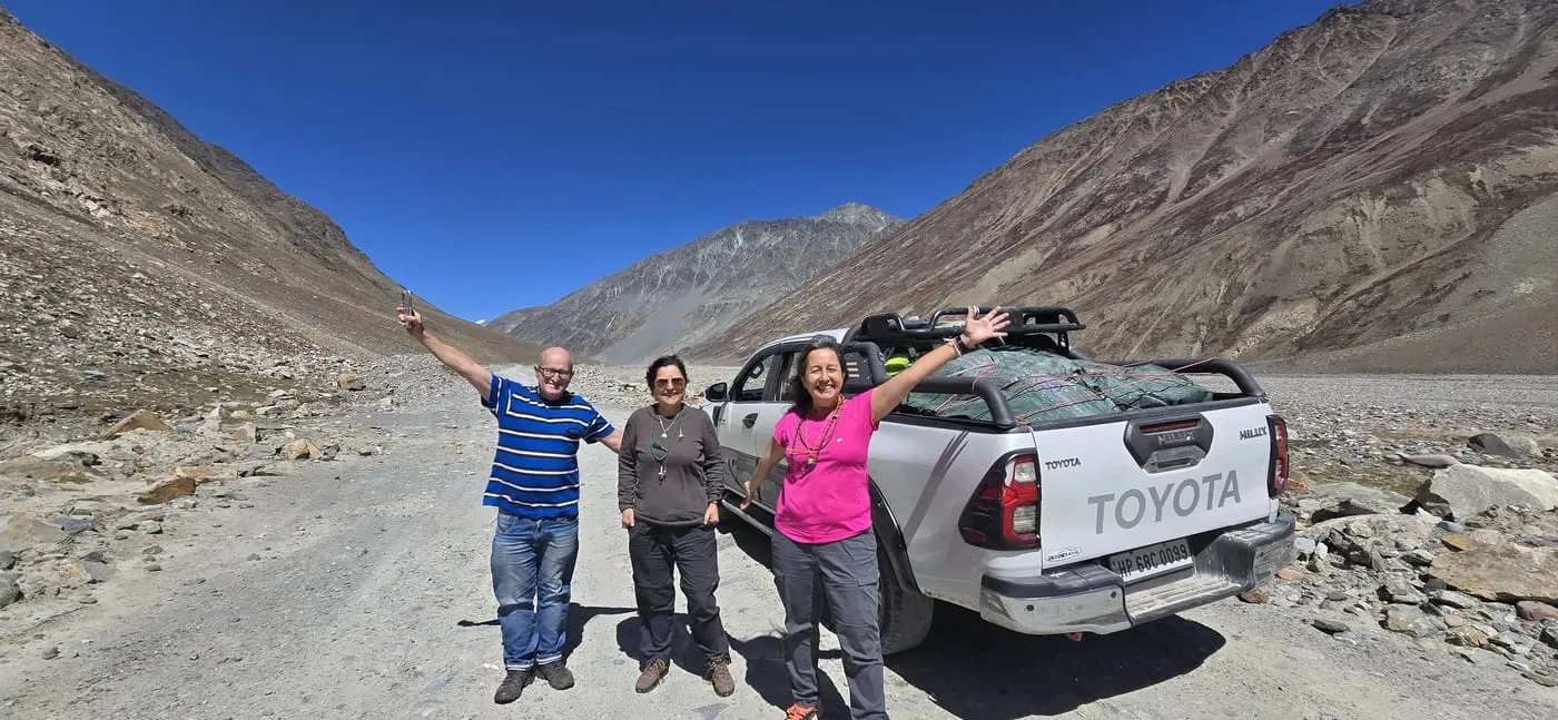 Three smiling travellers wave beside a white pickup on a gravel road between bare mountains in Spiti