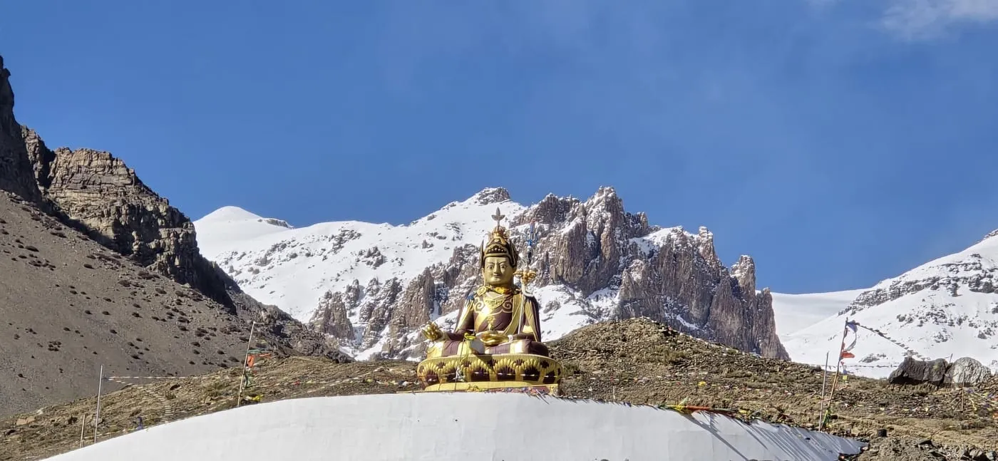The golden Buddha statue at Langza, sitting on a white base below snowy peaks and a blue sky