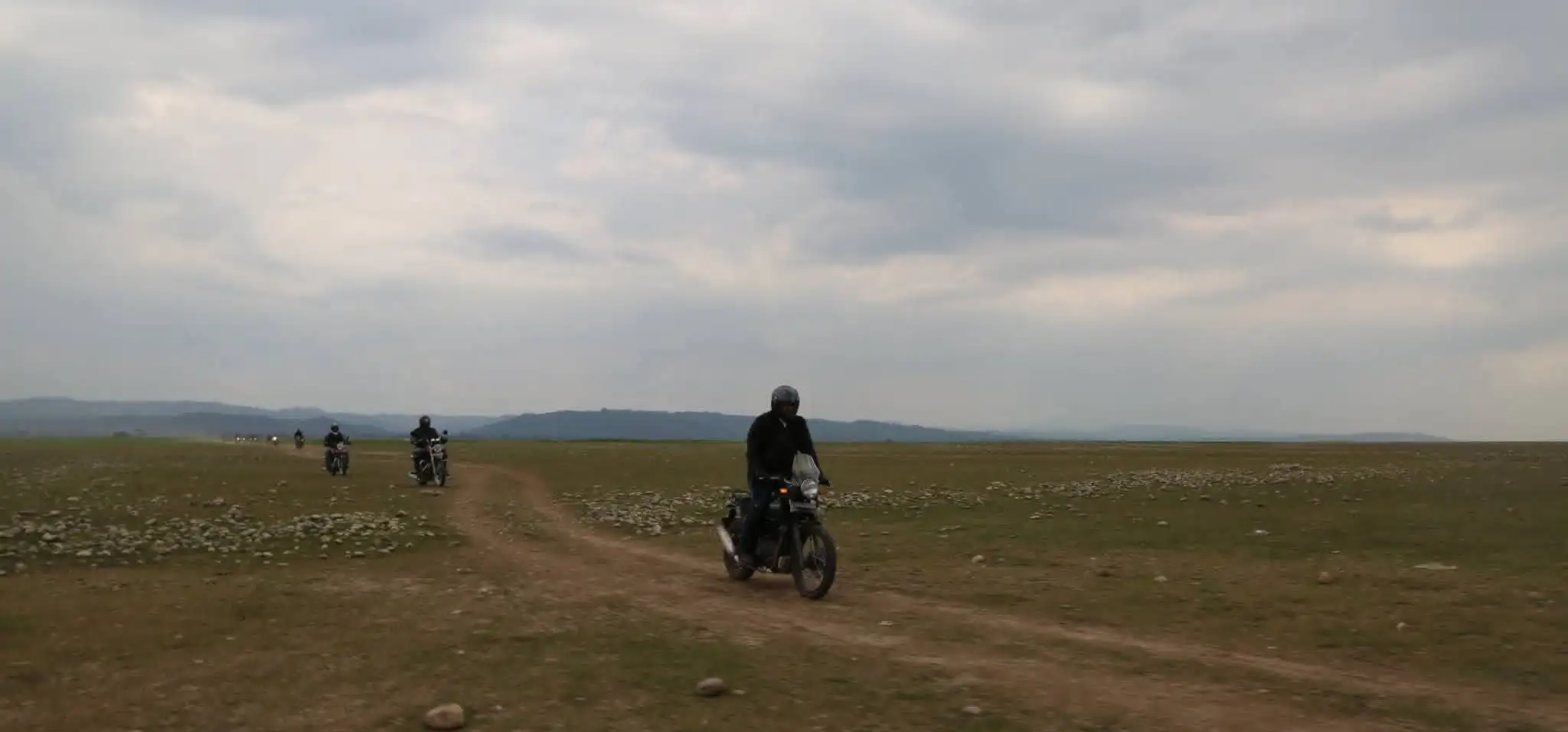 A line of riders on Royal Enfield motorcycles follows a dirt track across open grassland under a grey sky