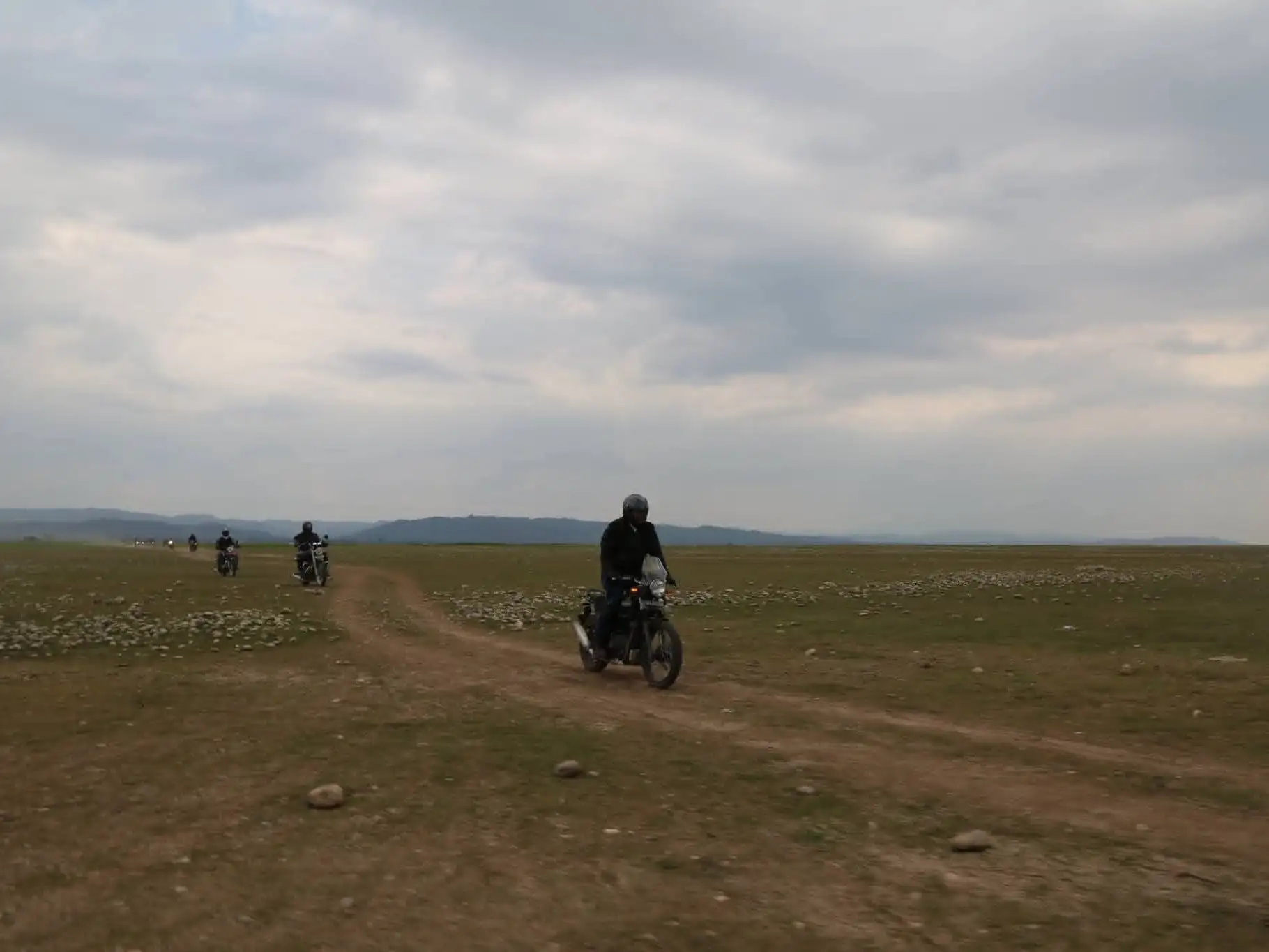 A line of riders on Royal Enfield motorcycles follows a dirt track across open grassland under a grey sky