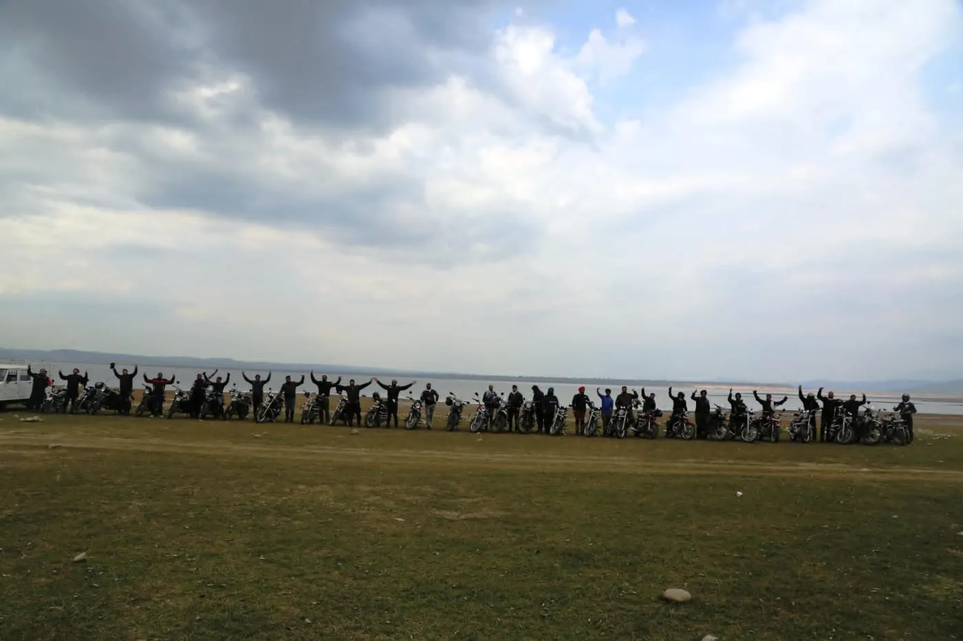 Riders and their Royal Enfields lined up with arms raised beside a wide lake under a cloudy sky