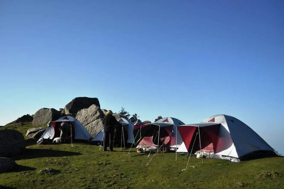 Tents pitched among granite boulders on a grassy Dhauladhar ridge under a clear blue sky