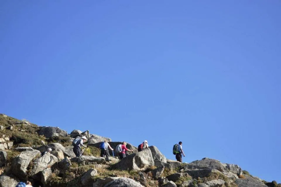A line of trekkers climbs a ridge of grey boulders against a clear blue sky