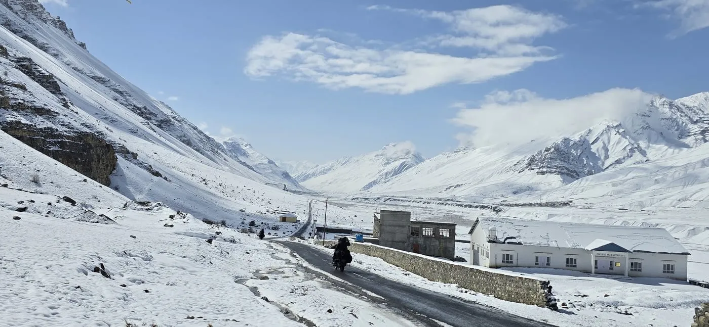 A motorcyclist rides a cleared road through snow near Kaza, with white mountains and a blue sky
