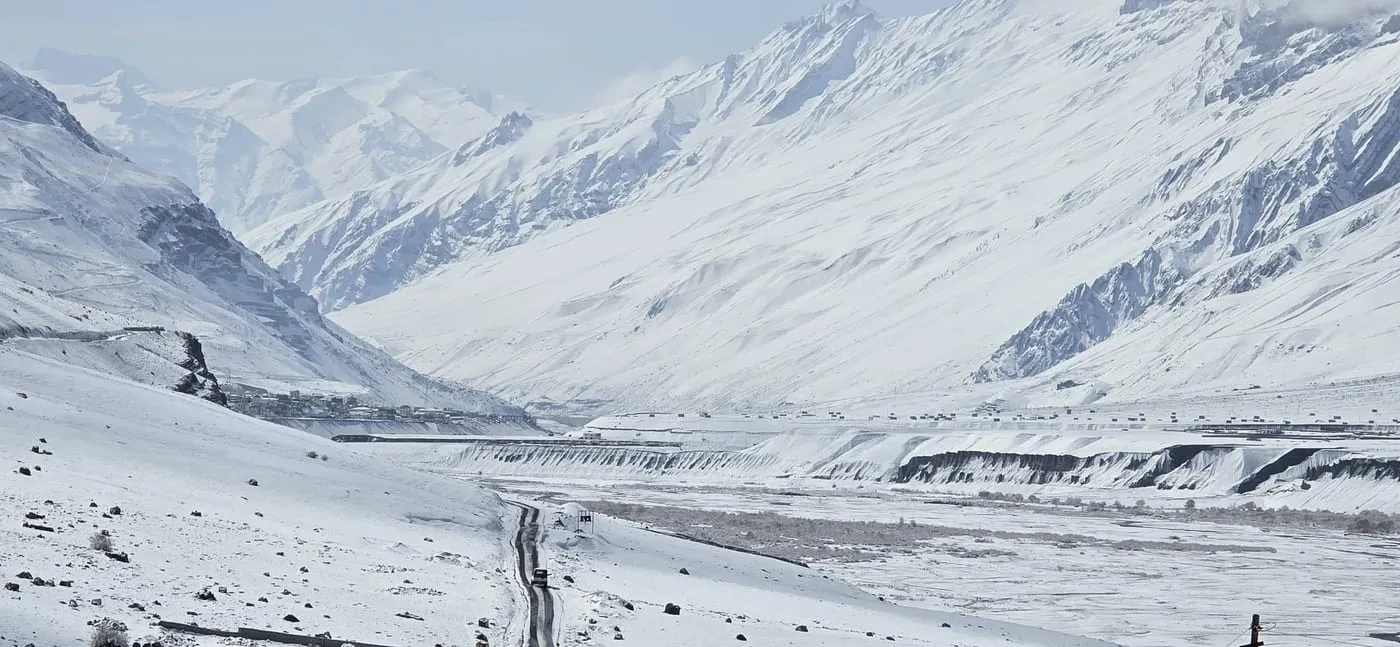 A car drives a narrow cleared road through a white valley in Spiti, with Kaza across the frozen river