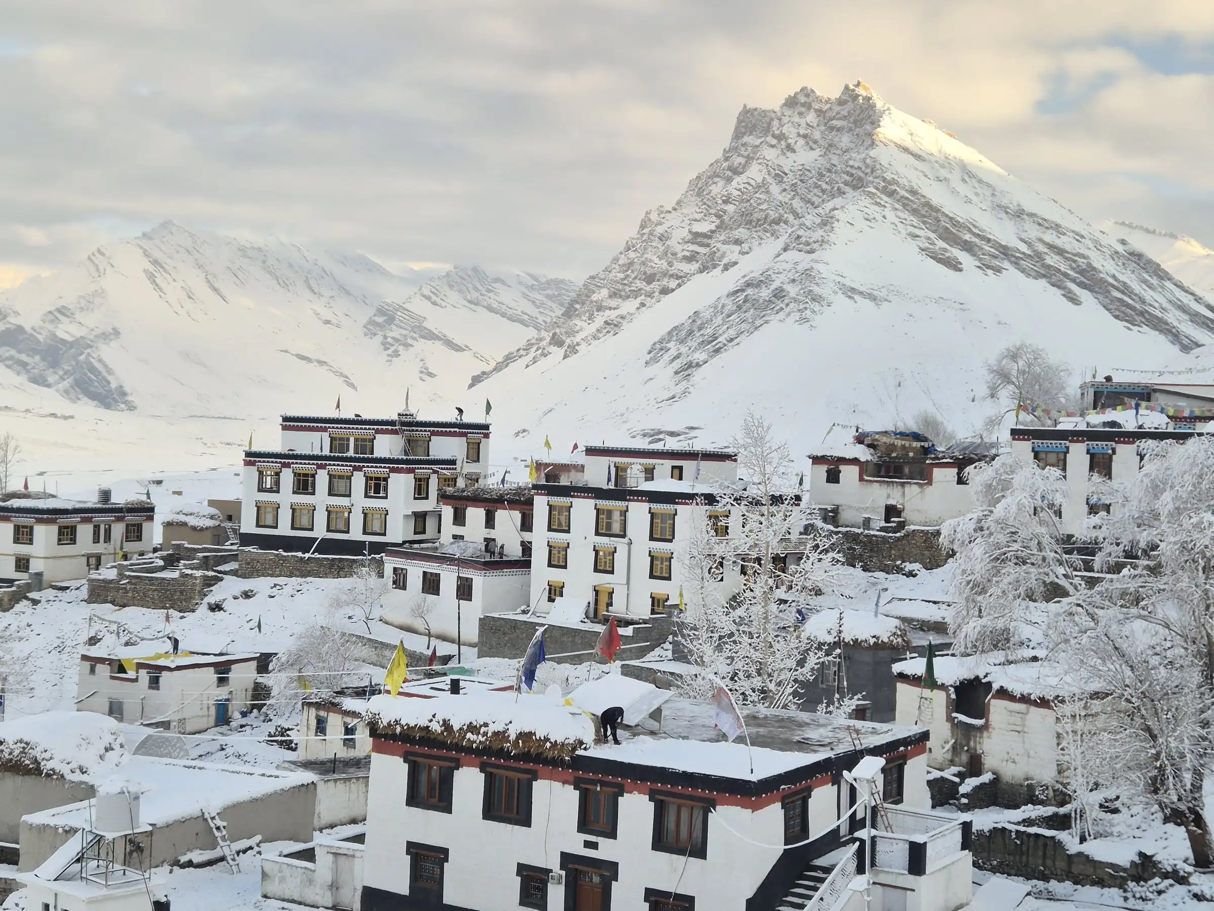 White houses and prayer flags under fresh snow in a Spiti village, with a snow-covered peak behind