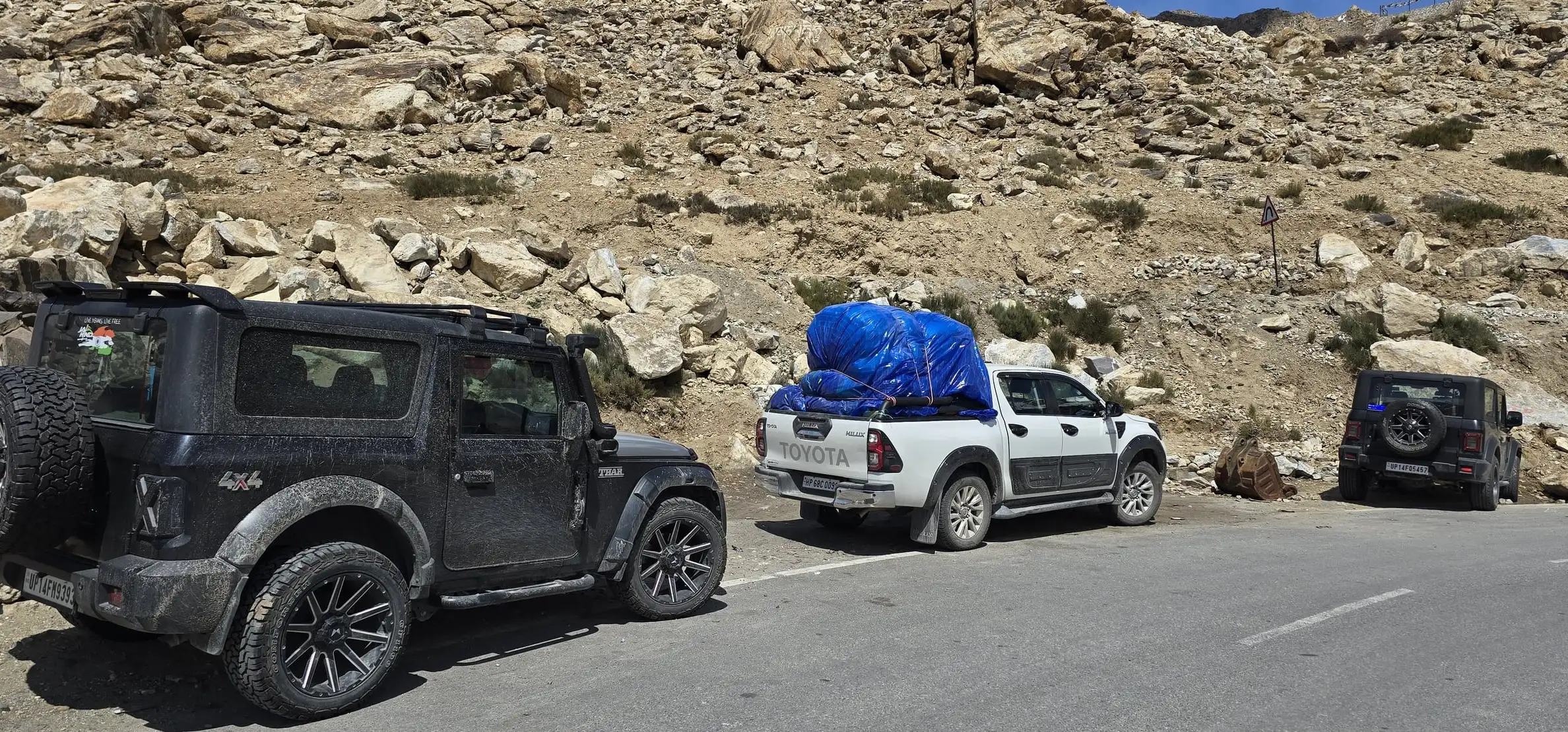 A black 4x4 and a white pickup loaded with luggage parked on a mountain road below rocky slopes