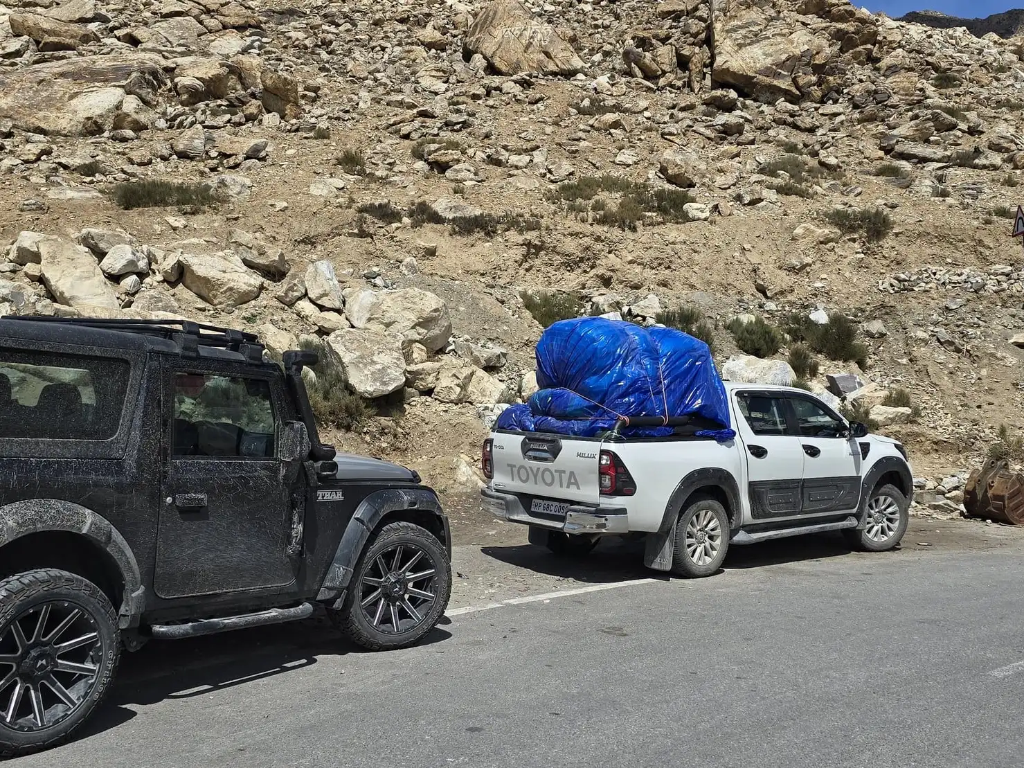 A black 4x4 and a white pickup loaded with luggage parked on a mountain road below rocky slopes